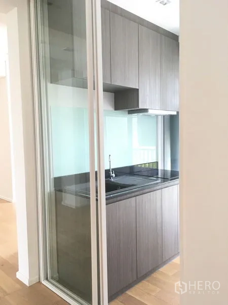 Condominium for rent or sale in Chatuchak, Bangkok - Compact enclosed kitchen behind a glass sliding door with grey cabinets and black countertop.