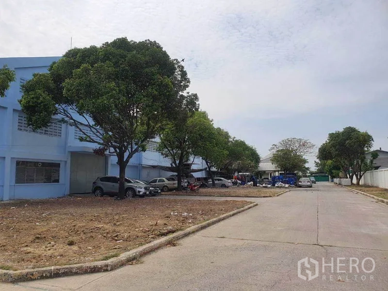 Warehouse for rent in Nakhon Pathom - Access road in the compound with trees and on-site parking beside warehouse blocks.
