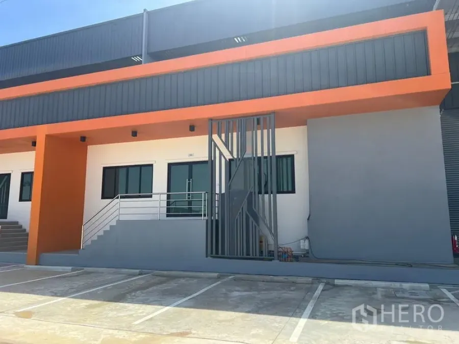 工厂 出租在Bang Pakong, Chachoengsao - 现代厂房外立面，橙色点缀，办公室窗户、楼梯与划线停车位。