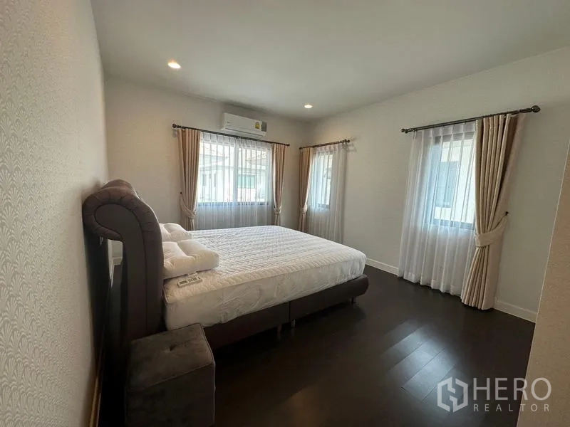 Detached House for rent or sale in Lam Luk Ka, Pathum Thani - Guest bedroom with double bed, three windows and curtains over dark wood flooring.