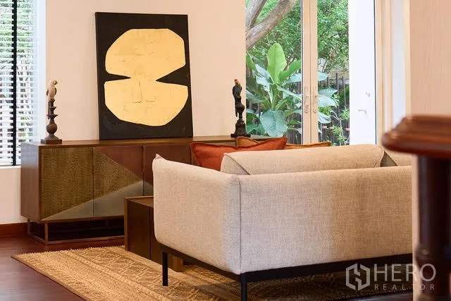 Detached House for rent in Watthana, Bangkok - Cozy seating corner with light sofa, cabinet and abstract artwork near garden doors.