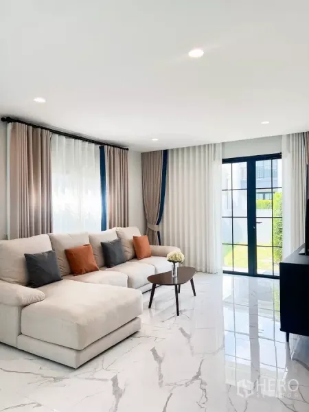 Detached House for rent in Khan Na Yao, Bangkok - Bright living room with large beige sectional sofa, marble-look floor tiles, and glass door opening to the garden.