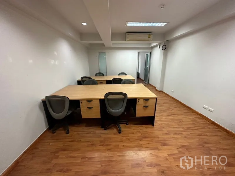 Office Space for rent in Khlong Toei, Bangkok - Open-plan workspace with multiple desks and chairs, wood laminate floor, ceiling lighting, and a wall-mounted air conditioner by a glass door.