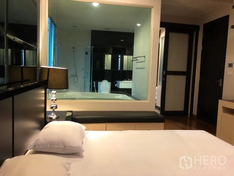 公寓 出租在Pathum Wan, Bangkok - 主卧带床头灯，透过玻璃可见带浴缸与淋浴间的套内卫浴。