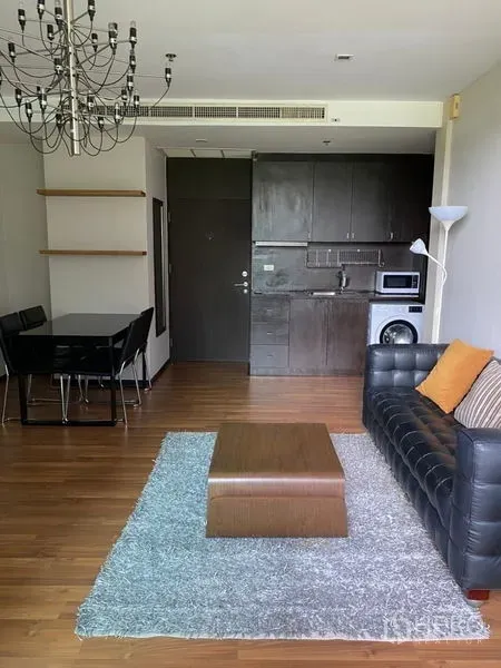 Condominium for rent in Khlong Toei, Bangkok - Open-plan living area with wood floors, dark built-in kitchenette with microwave and washer, dining set and leather sofa under a chandelier.