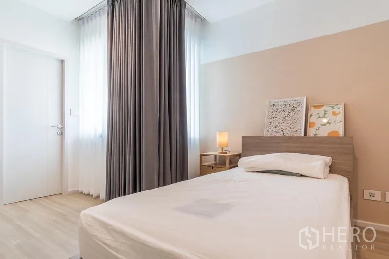 Townhouse for rent or sale in Suan Luang, Bangkok - Cozy secondary bedroom with single bed, bedside table, long drapes, and light wood flooring.