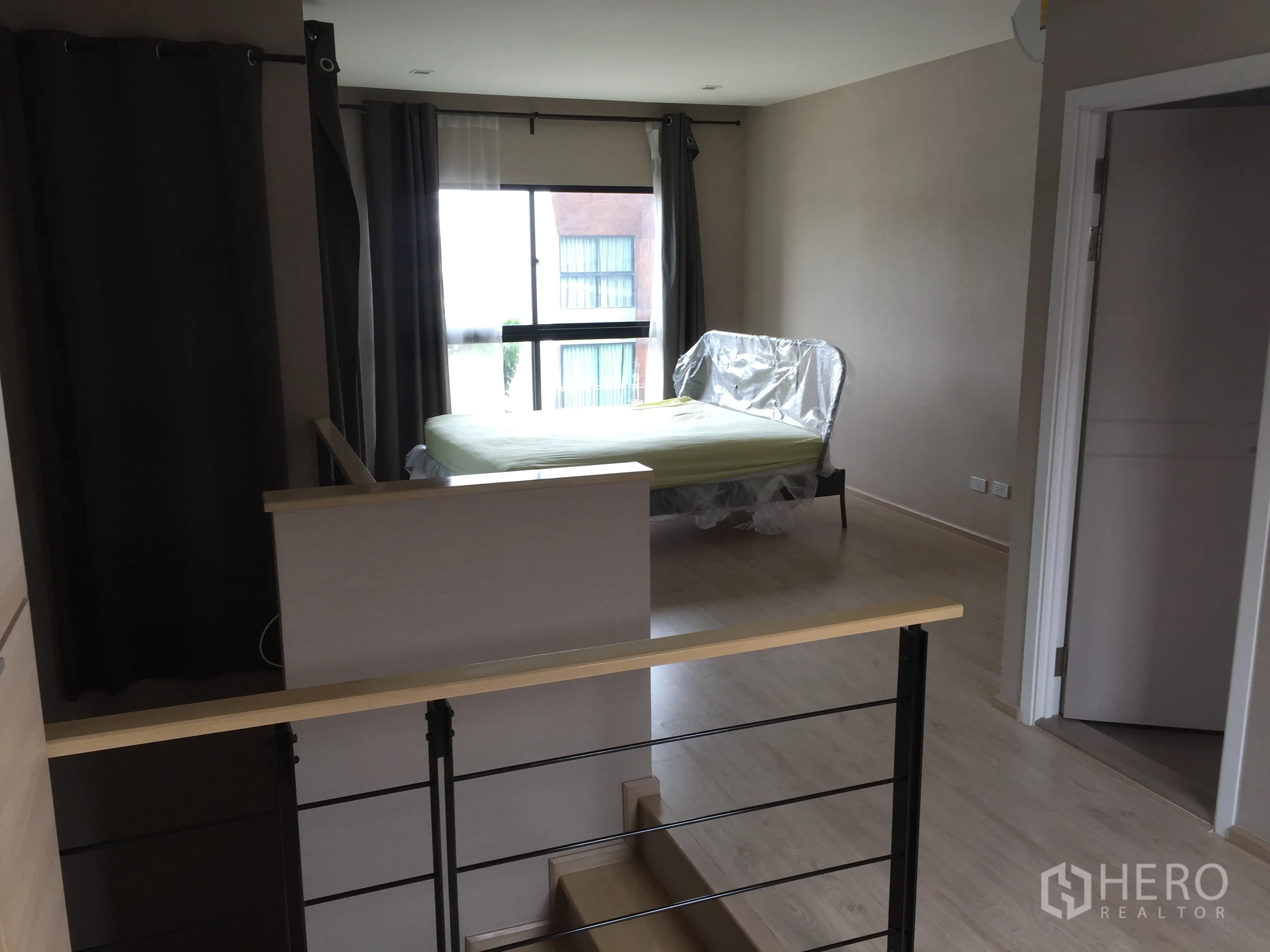 Townhouse for rent in Suan Luang, Bangkok - Master bedroom with large window, bed frame, curtains and doorway to bathroom near stair landing.