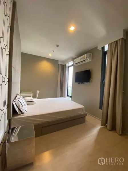 Condominium for rent in Watthana, Bangkok - Corner bedroom with wall-mounted TV, air conditioner and tall windows with curtains.