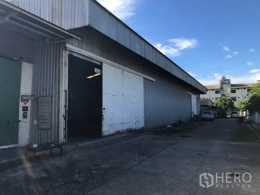 Warehouse for rent in Mueang Samut Sakhon, Samut Sakhon - Steel-clad warehouse exterior with large sliding door and driveway under blue sky.
