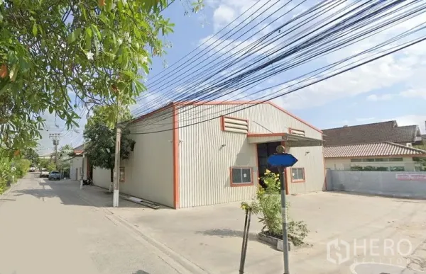 Warehouse for rent in Mueang Nonthaburi, Nonthaburi - Street view of a corrugated metal warehouse with gable roof, roller shutter entrance and wide concrete driveway on a quiet lane in Bang Kraso, Nonthaburi.