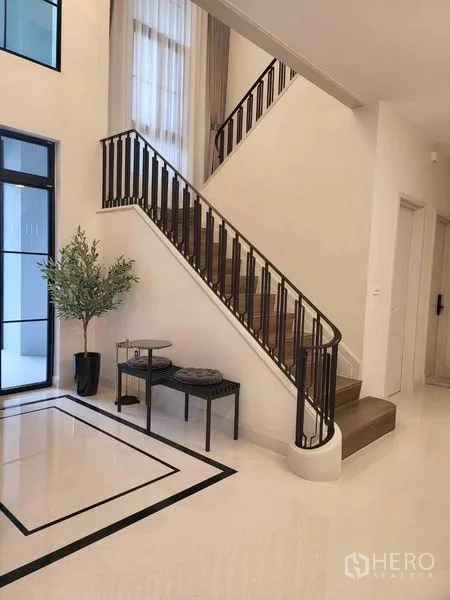 Detached House for rent in Saphan Sung, Bangkok - Staircase with dark handrail and a tall window in a bright foyer.