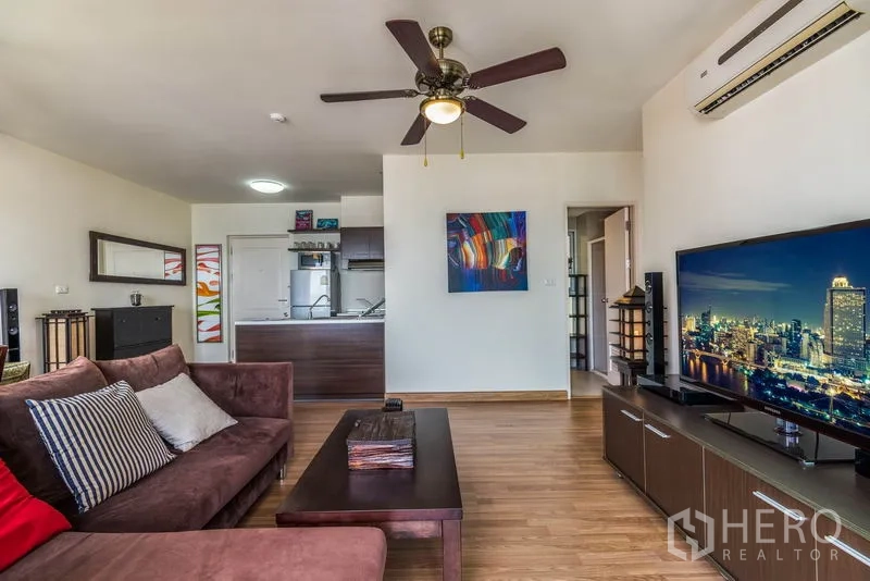 Condominium for rent in Bang Na, Bangkok - Living room with TV console, ceiling fan, and view toward the open-plan kitchen.