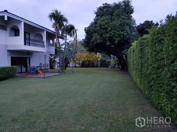 Detached House for rent in Khan Na Yao, Bangkok - Expansive private garden with manicured lawn, tall hedge and balcony view of a two-storey home.