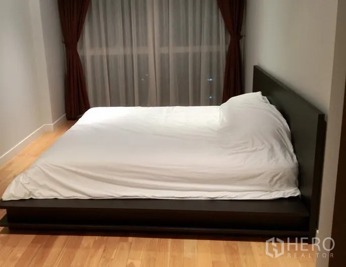 Condominium for rent in Khlong Toei, Bangkok - Quiet bedroom with a low platform bed, wooden floors, and full-height windows with dark curtains.