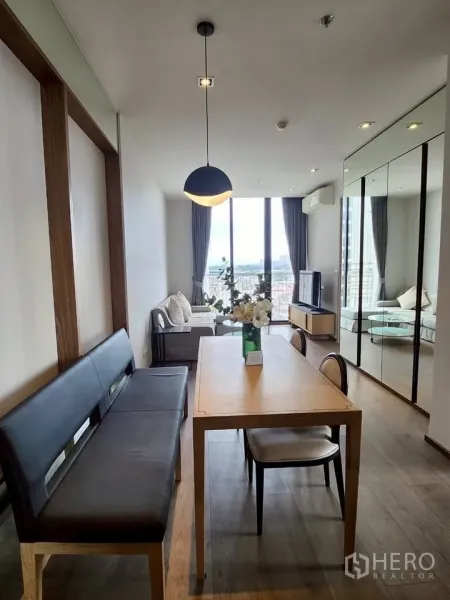 Condominium for rent in Khlong Toei, Bangkok - Dining table with built‑in bench leading to a bright living room with floor‑to‑ceiling windows and city view.