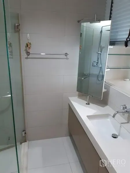 Condominium for rent in Khlong Toei, Bangkok - Clean bathroom with a glass-enclosed shower, white vanity, wall mirror, and light tiled walls.