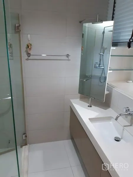 Condominium for rent in Khlong Toei, Bangkok - Clean bathroom with a glass-enclosed shower, white vanity, wall mirror, and light tiled walls.