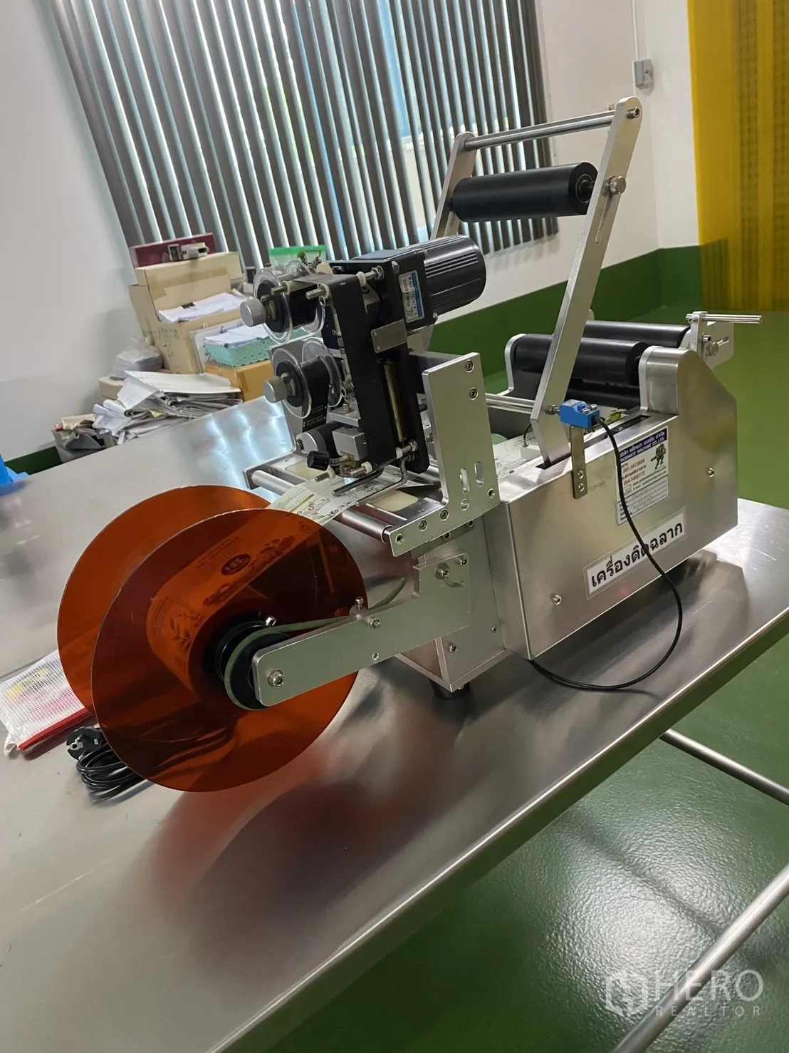 Factory for sale in Khlong Luang, Pathum Thani - Labeling machine on a stainless worktable beside window blinds.