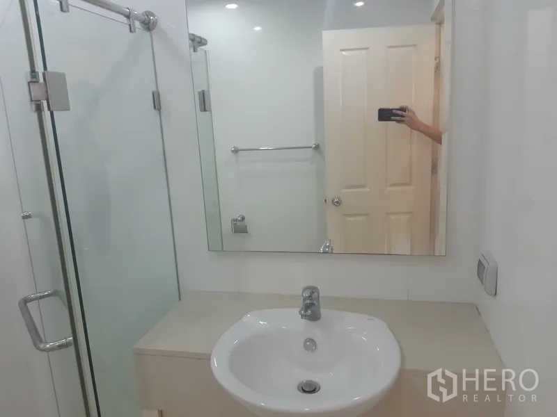 Condominium for rent in Khlong Toei, Bangkok - Compact bathroom vanity with wide mirror beside a clear glass shower enclosure.