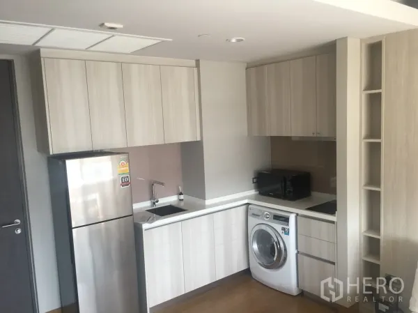 Condominium for rent in Khlong Toei, Bangkok - Modern compact kitchen with light wood cabinets, fridge, microwave, cooktop, and washing machine.