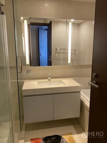 Condominium for rent in Watthana, Bangkok - Contemporary bathroom with lighted mirror cabinet, white vanity and partial bathtub in neutral tiles.