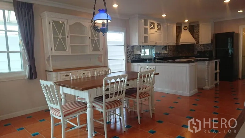 Detached House for rent in Mueang Samut Prakan, Samut Prakan - Dining area beside an open kitchen with white cabinetry, island, terracotta floor tiles and pendant light.