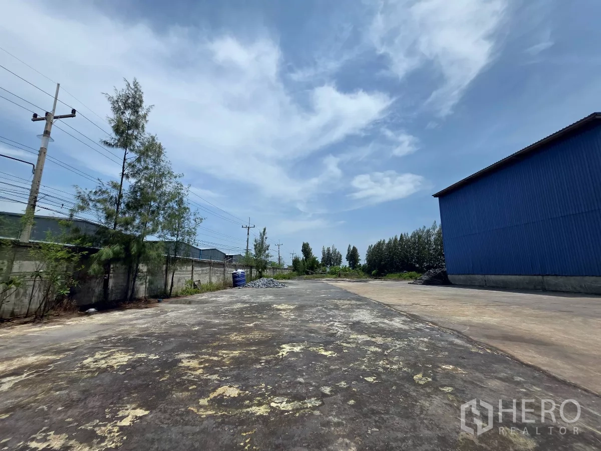 โรงงาน ให้เช่าในMueang Samut Sakhon, Samut Sakhon - ลานรอบเขตที่มีกำแพงรั้ว สายไฟฟ้า และต้นไม้ ข้างโกดังสีน้ำเงิน