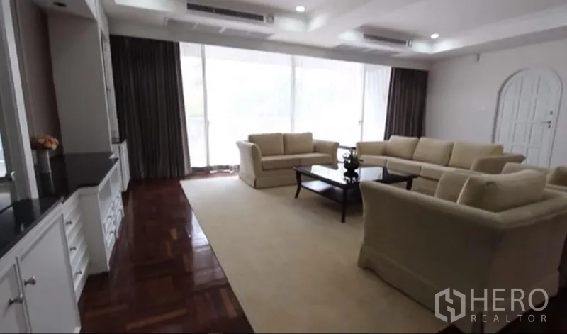 房间 出租在Khlong Toei, Bangkok - 温馨客厅一角，米色沙发、茶几与整面窗帘玻璃。
