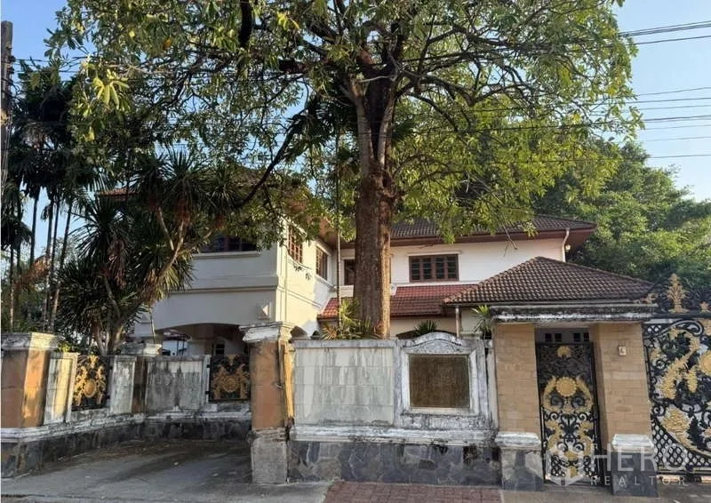 Detached House for sale in Sai Mai, Bangkok - Close view of the main gate and driveway showing an arched porch, upper balcony and a big shade tree at a two-storey home.