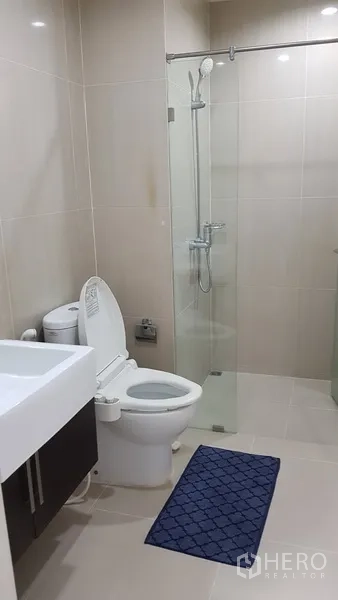 Condominium for rent in Ratchathewi, Bangkok - Second bathroom with glass-enclosed shower, toilet and vanity in beige tiles.