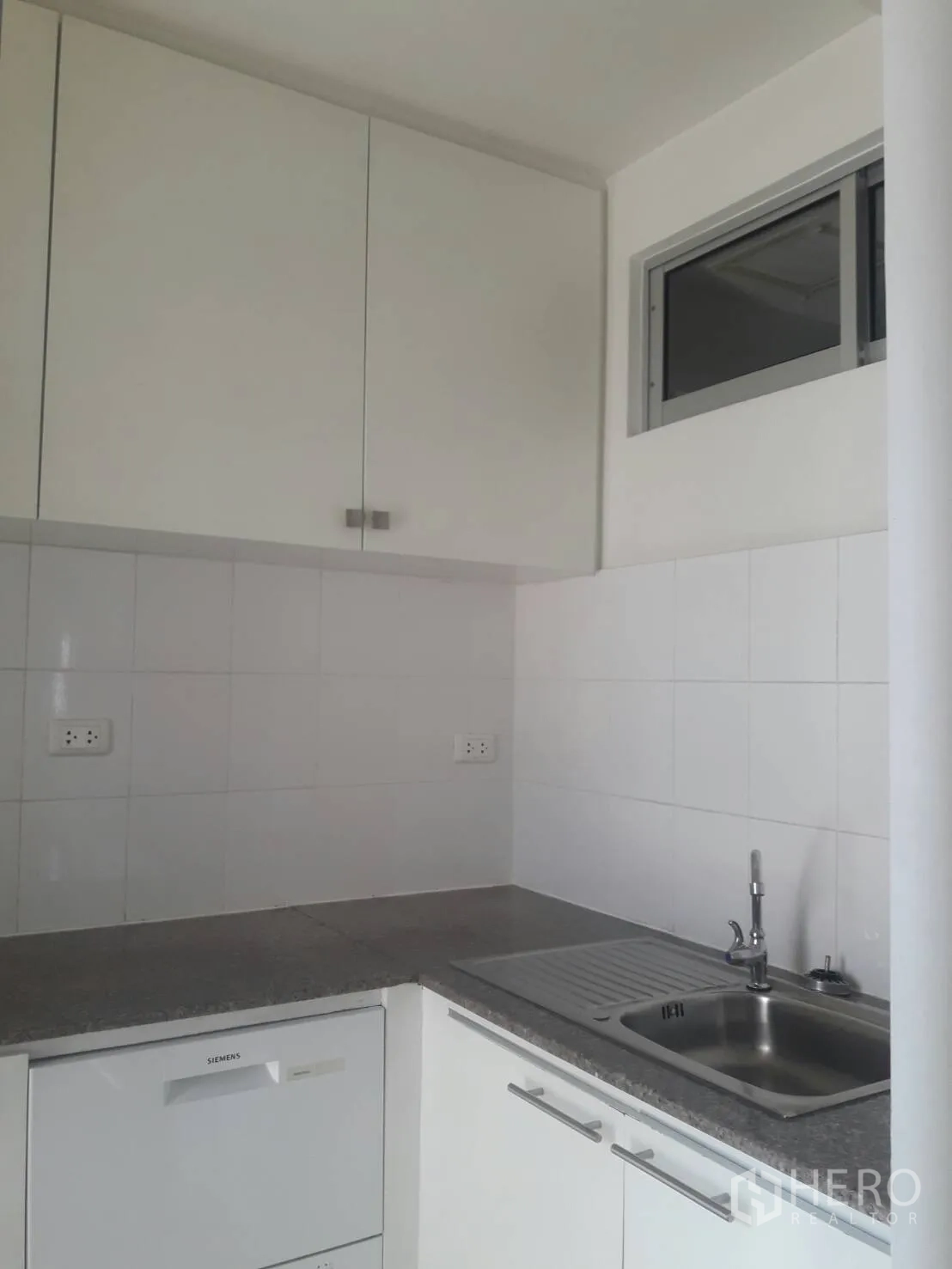 Condominium for rent in Khlong Toei, Bangkok - Compact white L-shaped kitchen with tiled walls, upper cabinets, stainless sink and a small sliding window.