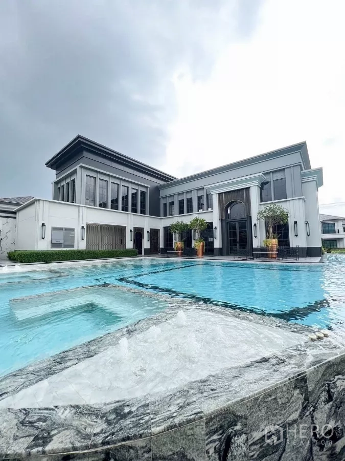 Detached House for rent in Khan Na Yao, Bangkok - Estate clubhouse with large swimming pool and modern architecture under cloudy sky.