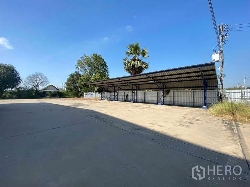 货栈 出租在Lat Lum Kaeo, Pathum Thani - 有顶停车位，位于宽阔硬化场地，背景有围栏与树木。