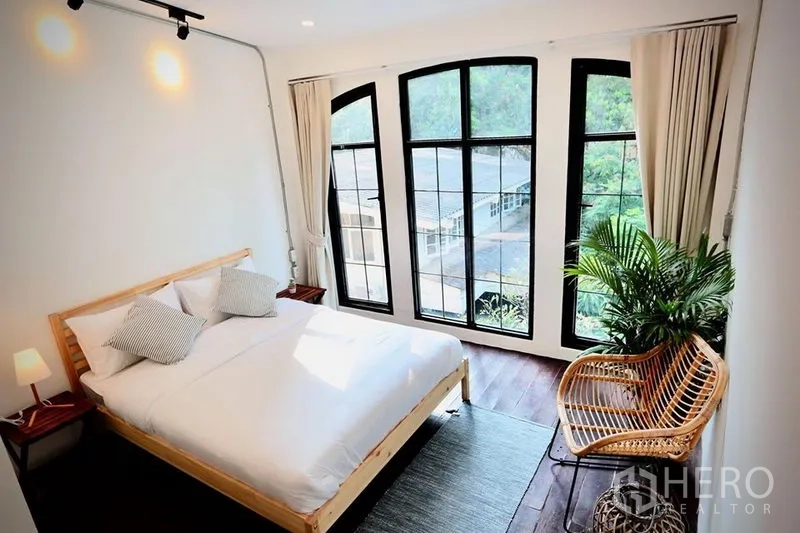 Office Space for rent in Sathon, Bangkok - Sunlit bedroom with large arched black-framed windows, wooden bed and rattan chair.