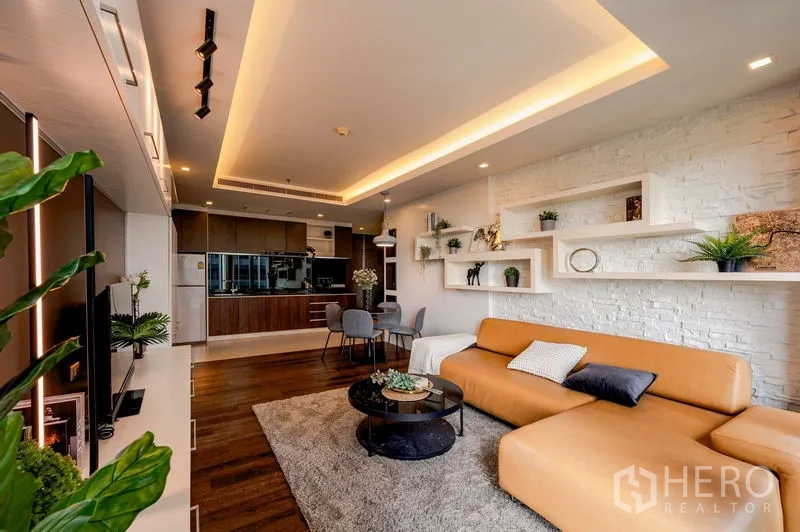 Condominium for sale in Khlong Toei, Bangkok - Living area with white brick accent wall, floating shelves, tan sofa, dining set, and kitchen in the background.