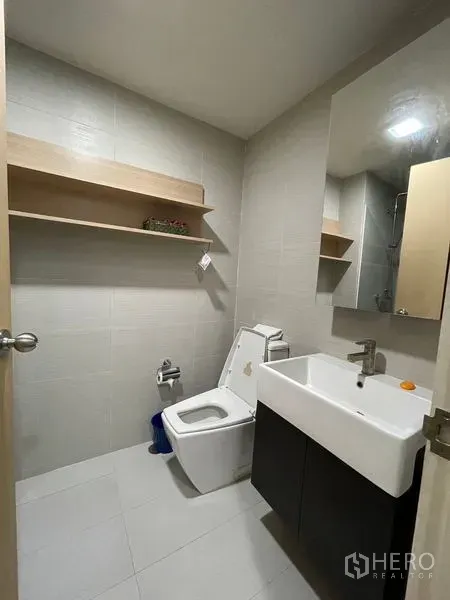 Condominium for sale in Khlong Toei, Bangkok - Clean bathroom with large mirror, vanity sink, toilet and a floating wall shelf.