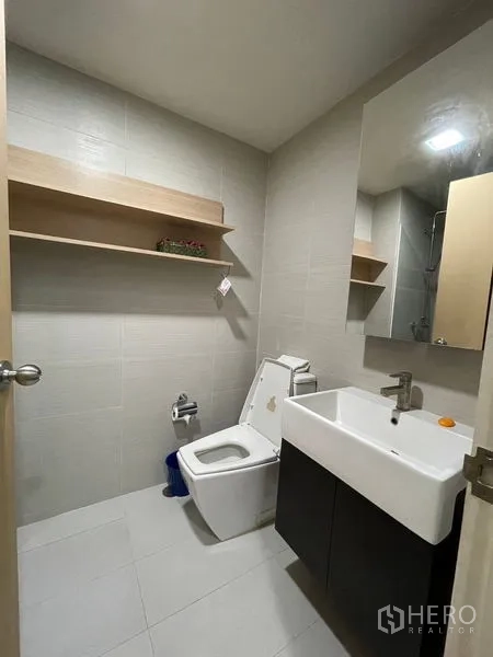 Condominium for sale in Khlong Toei, Bangkok - Clean bathroom with large mirror, vanity sink, toilet and a floating wall shelf.