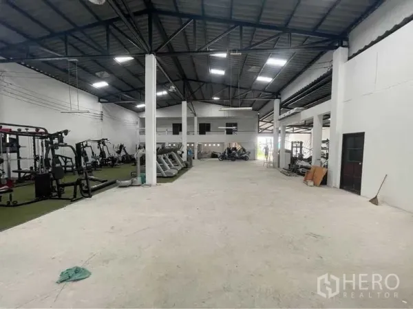 Warehouse for sale in Lam Luk Ka, Pathum Thani - Open-plan warehouse hall with steel roof trusses, bright skylights and fitness machines lined along one side.