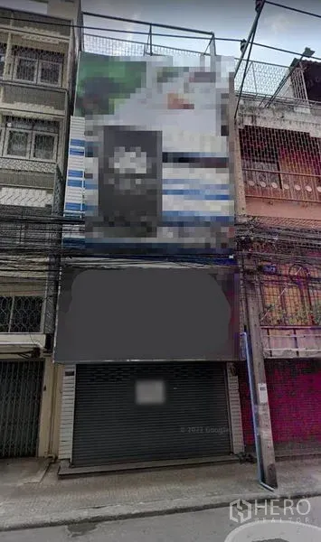 Shophouse for rent in Khlong Toei, Bangkok - Multi‑storey shophouse facade on a narrow Bangkok street with metal roller shutter, blank signage area, and overhead utility cables.