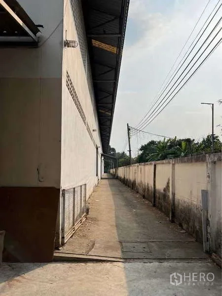 คลังสินค้า ขายในBang Bua Thong, Nonthaburi - ทางเดินคอนกรีตแคบขนานตัวอาคารพร้อมแนวรั้วและสายสาธารณูปโภค.