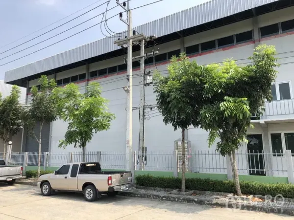 Factory for rent in Lam Luk Ka, Pathum Thani - Side facade of the factory showing high windows, perimeter fence, trees and roadside parking.