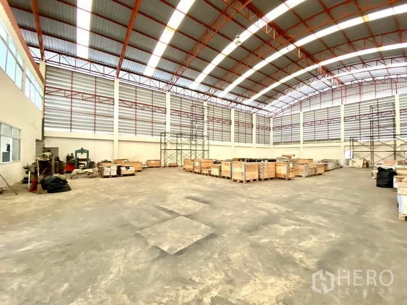 Warehouse for rent in Lam Luk Ka, Pathum Thani - Wide storage bay with steel trusses, pallets and racking frames along the walls.