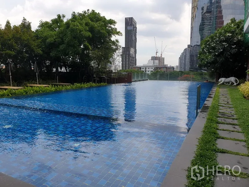 Condominium for rent or sale in Watthana, Bangkok - Outdoor swimming pool with blue tiles, landscaped deck and city skyline backdrop.