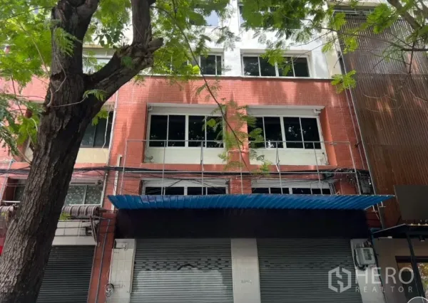 店屋 出租在Bang Rak, Bangkok - 邦拉克四层店屋外观，红色瓷砖立面，大窗与蓝色雨棚，街道树荫。