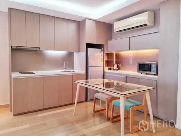 Condominium for rent in Watthana, Bangkok - Fitted kitchen with light wood cabinetry, built-in appliances and glass dining table with stools.