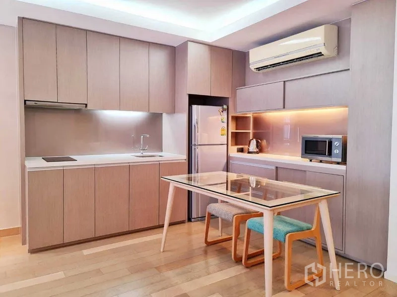 Condominium for rent in Watthana, Bangkok - Fitted kitchen with light wood cabinetry, built-in appliances and glass dining table with stools.