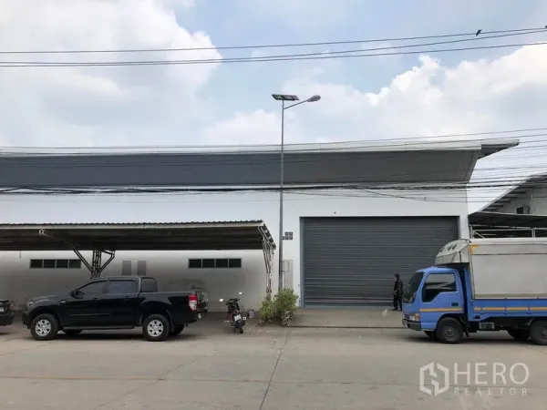 Factory for rent in Nonthaburi - Street view of a white factory with a large roller shutter, canopy parking and delivery trucks in front.