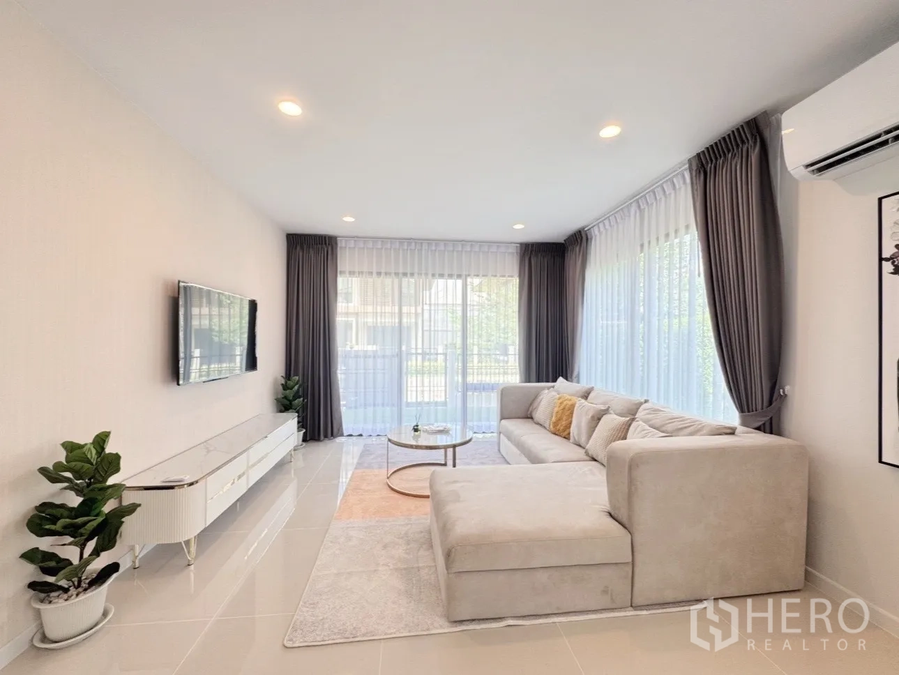 Detached House for rent or sale in Bang Kapi, Bangkok - Living room with large windows, grey curtains, wall-mounted TV and sectional sofa.