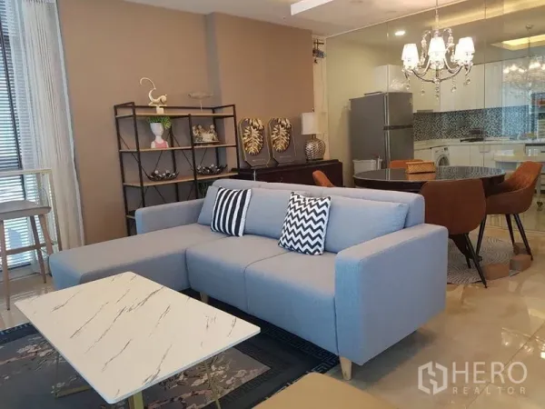 Condominium for rent or sale in Bang Na, Bangkok - Open-plan living and dining area with a light gray sectional sofa, marble coffee table, crystal chandelier, and a glass-partitioned kitchen.