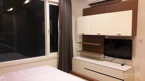 Condominium for rent in Khlong Toei, Bangkok - Bedroom wall with mounted cabinets, open shelves, flat-screen TV and a wide window with curtains and AC.